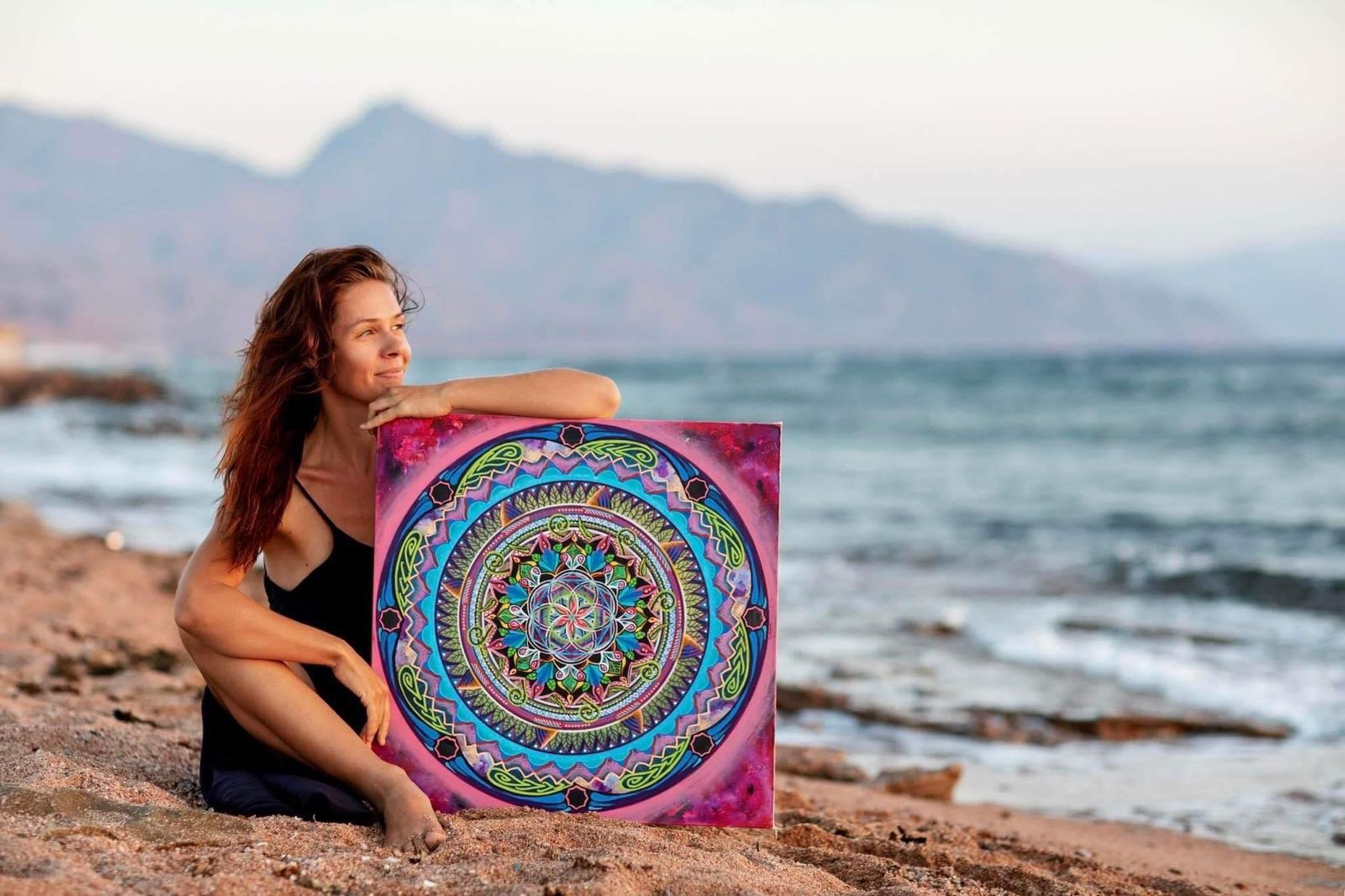 Ela sitting on a rocky beach at sunset with a vibrant mandala painting, ocean and mountains behind her